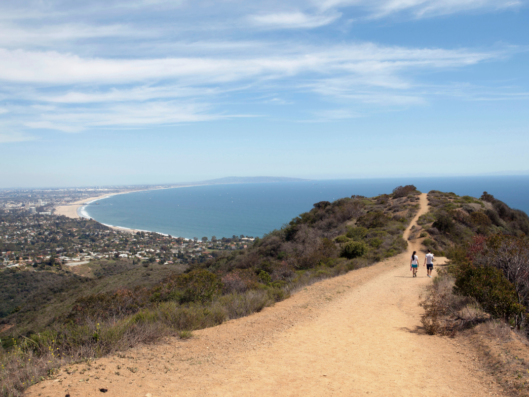 A photo of two people walking down Los Liones Trail with a view of the ocean.