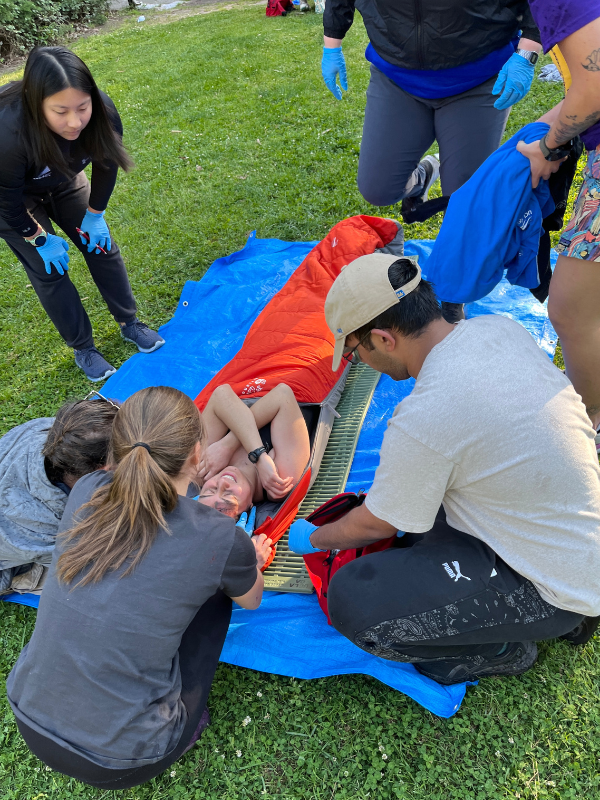 A group of people practice wrapping and securing a person on a tarp with a sleeping bag during an outdoor emergency response training.