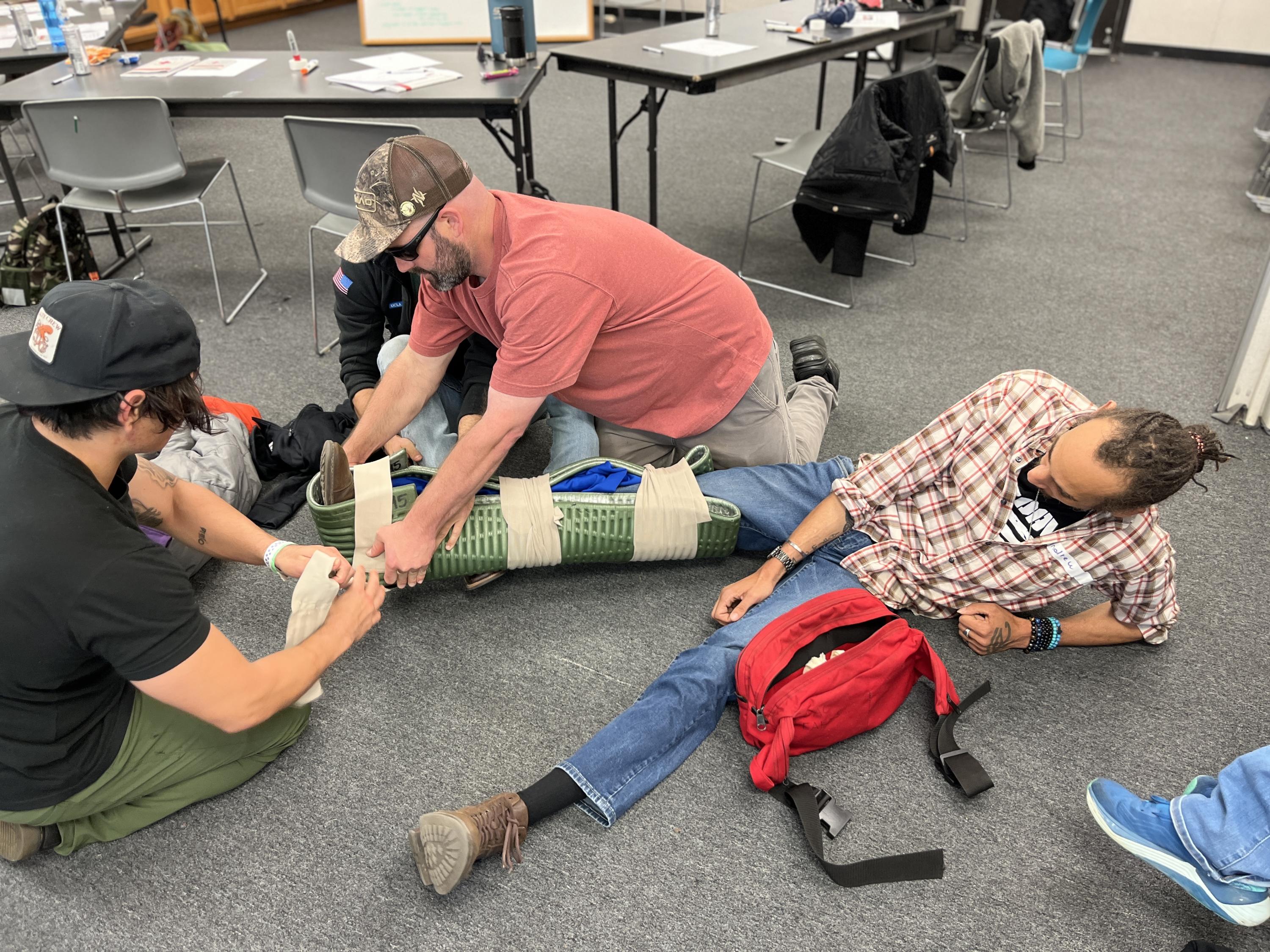 Three people practice a first aid drill indoors, using a foam mat and bandages to splint a man's leg. The setting appears to be a classroom or training space, with chairs and supplies nearby.